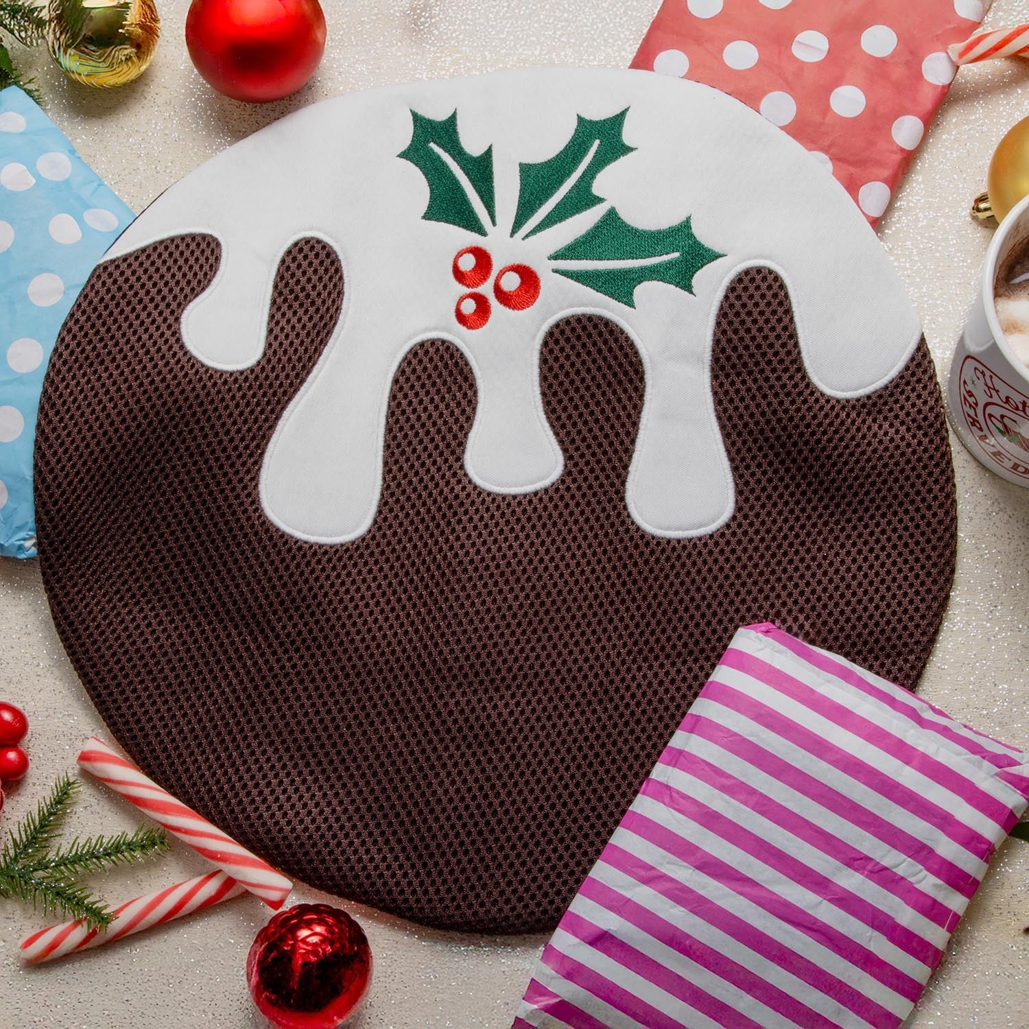 Une photo flat lay festive pour le calendrier de l'Avent Snagvent 2025, centrée sur un grand sac de toilette rond conçu pour ressembler à un pudding de Noël. Le sac est composé d'un corps en maille brune, d'un "glaçage" en tissu blanc qui coule sur le dessus, et de feuilles de houx vertes brodées avec des baies rouges. Il est placé sur une surface pailletée, entouré d'objets de Noël, notamment des sacs cadeaux en papier coloré, des sucres d'orge, des boules rouges et dorées et une tasse de chocolat chaud.