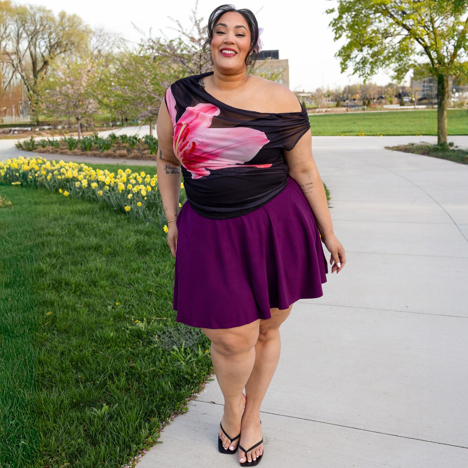 Mujer con un top floral de un solo hombro y una falda morada, de pie al aire libre con vegetación y flores al fondo.