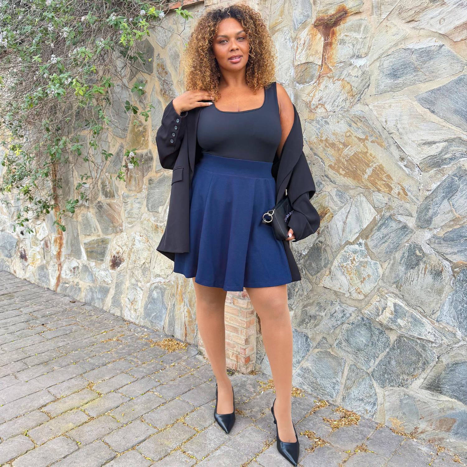 Una mujer con un vestido azul marino y una chaqueta negra, de pie junto a un muro de piedra.