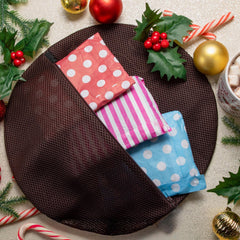 Le verso du sac de lavage Snagvent Christmas pudding, montrant le tissu en maille marron et l'ouverture à fermeture éclair. À l'intérieur se trouvent trois paires de collants dans un emballage en papier coloré - rouge à pois blancs, rayures roses et blanches, et bleu à pois blancs - sur un fond festif de houx et de boules.